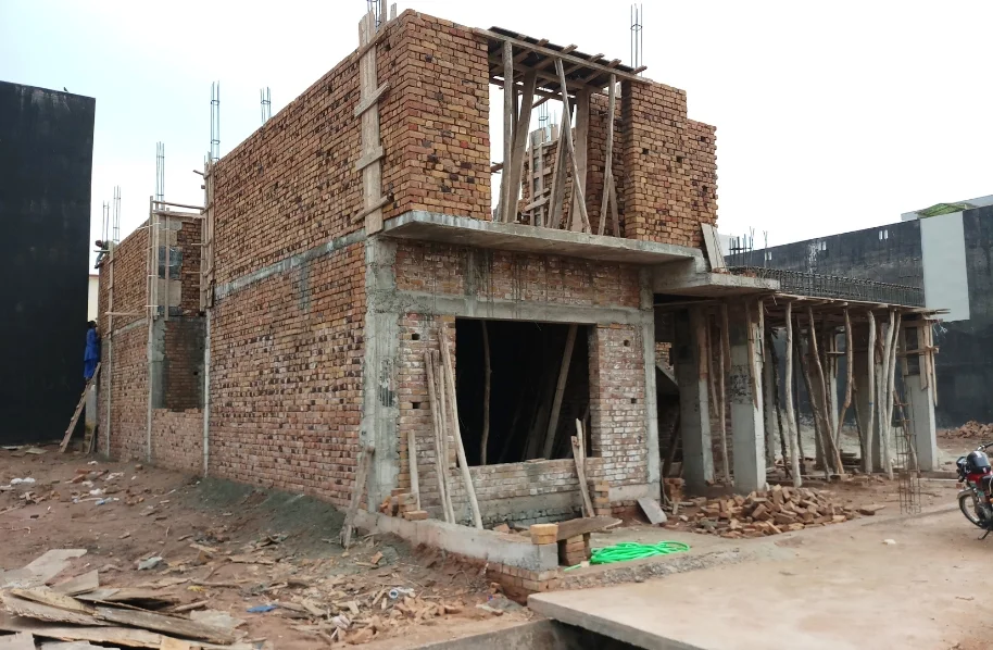 Construction workers and stacks of red bricks next to the partially built walls of the ground floor structure.