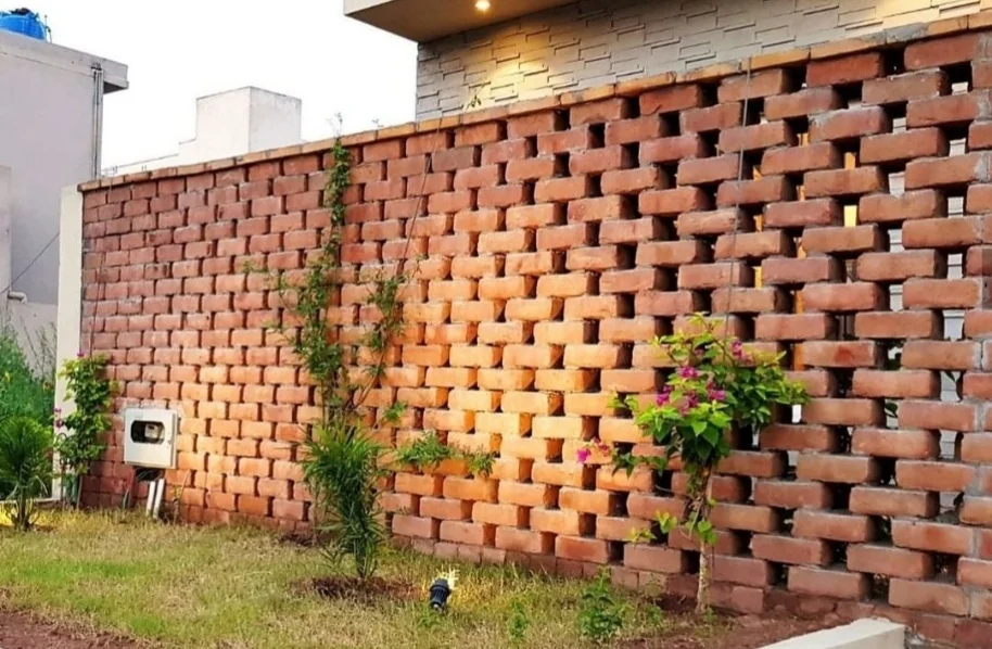 Close-up of a decorative red brick Jali (perforated screen) wall next to a small planted tree on a home exterior.